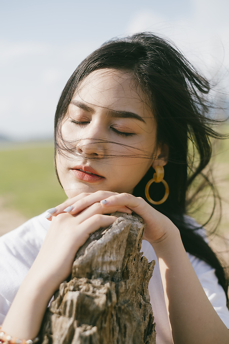 Black long hair woman in casual clothes prop her hands and head on the dry log, close her eyes in windy day.