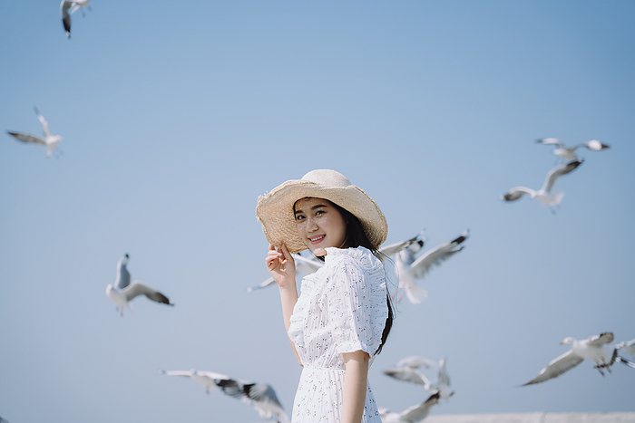 Long hair woman in white dress and hat standing in sunshine under blue sky with seagulls.