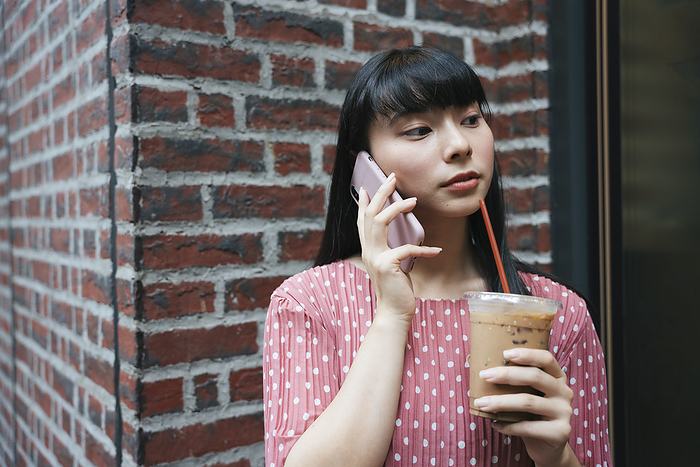 Beautiful asian thai long dark hair woman using smartphone while drinking ice coffee on street over brick background.