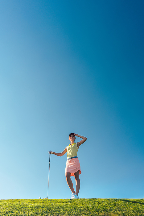 Fit and cheerful woman during practice on the green grass of a p Low angle view of a fit and cheerful woman wearing golf trendy outfits, while looking away during practice on the green grass of a professional golf course