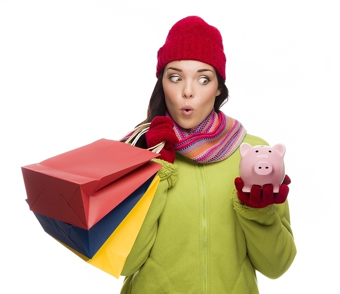 Concerned mixed-race woman wearing winter clothes holding shopping bags and piggybank isolated on white background, Photo by Andy Dean