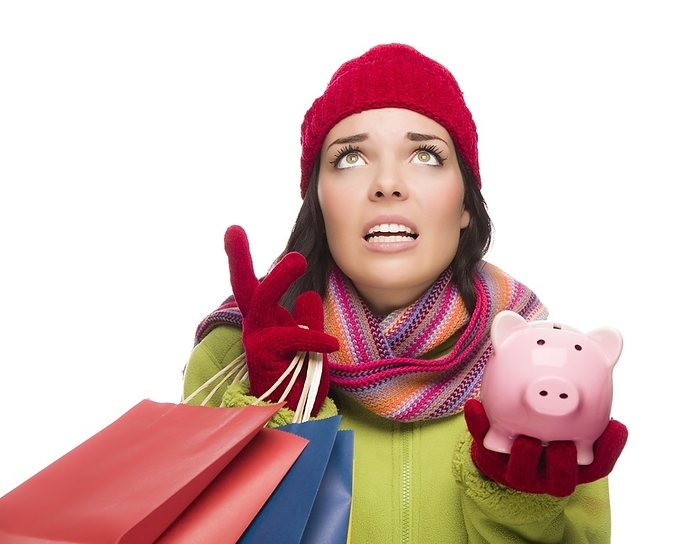 Stressed mixed-race woman wearing winter clothing looking up holding shopping bags and piggybank isolated on white background, Photo by Andy Dean