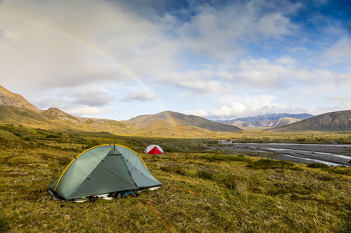 A rainbow arcs over a green backpacking tent and a red backpacking tent set up on the tundra with Marsh Fork River and mountains in the background,on a sunny summer day in the arctic; Brooks Range, Arctic National Wildlife Refuge (ANWR), Alaska, USA, Photo by Amber Johnson / Design Pics