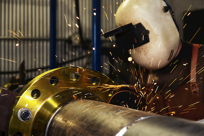 A welder wearing mask and eye protection is grinding a flange butt weld in a fabrication plant; Innisfail, Alberta, Canada, Photo by LJM Photo / Design Pics