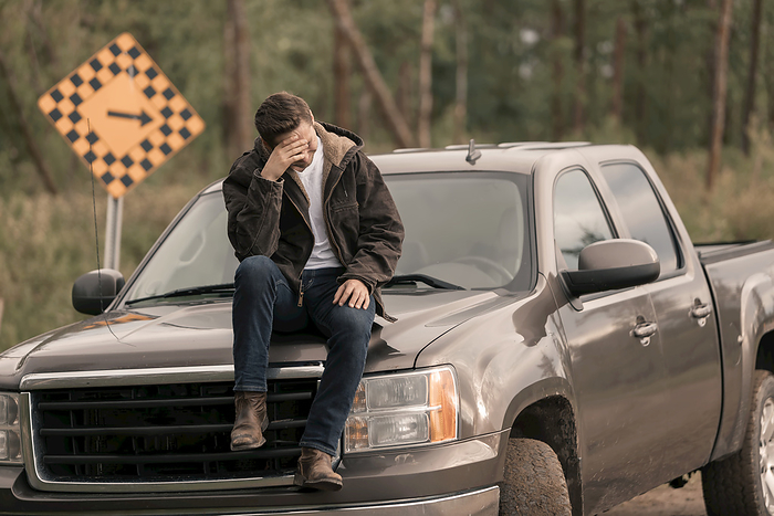 masculine gender A young man sits on a truck hood holding his head in his hand in distress, a directional roadside sign with arrow blurred in the background  Alberta, Canada, Photo by 770 Productions   Design Pics