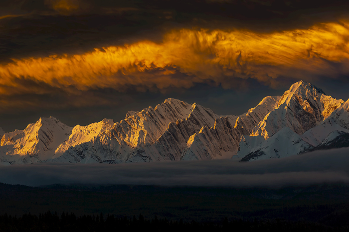 Canadian Rockies Dramatic clouds and snow covered mountain range in the winter at sunrise with warm light and highlights on clouds and mountains with fog in the valley, West of Calgary  Alberta, Canada, Photo by Michael Interisano   Design Pics