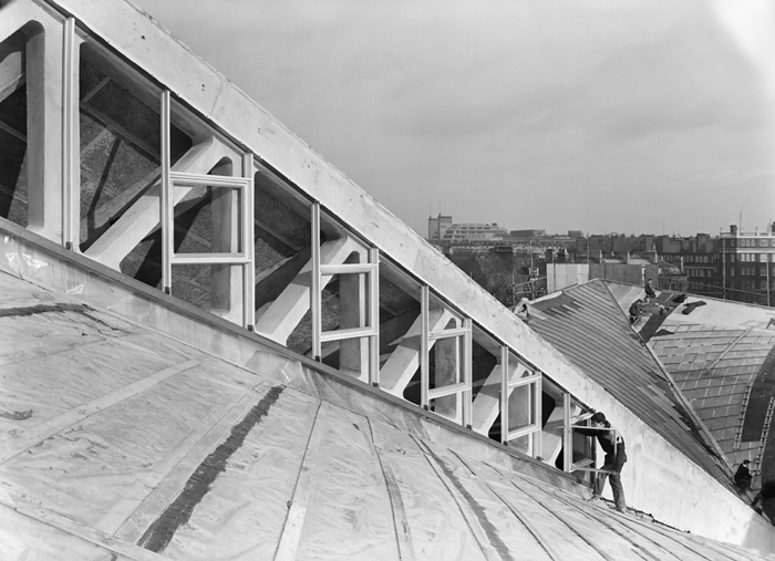 Commonwealth Institute, Kensington High Street, Kensington, London, 06 04 1962. Creator: John Laing plc. Commonwealth Institute, Kensington High Street, Kensington, London, 06 04 1962. A worker installing window frames in the clerestory between the central and outer roof sections of the exhibition hall at the Commonwealth Institute. Laing built the Commonwealth Institute between October 1960 and October 1962 to replace the former Imperial Institute that was to be demolished to make way for new facilities at Imperial College.  The building  consisted of a four storey administrative block housing a library, restaurant, board room and conference hall and a separate two storey block containing a cinema with an art gallery above, but the focus of the project was the exhibition hall with its hyperbolic paraboloid roof, the first of its kind constructed in Great Britain.  The exhibition, designed by James Gardner, provided spaces where each of the Commonwealth nations could showcase their achievements and characteristics, primarily to school children as teaching aids to enliven history and geography lessons. The shell arch of the central roof section was of reinforced concrete, cast in situ using timber formwork with rough sawn boards to provide a textured internal surface whilst the four outer  quot warps quot  were constructed using precast beams and wood wool slabs, blocks of shredded timber bound together in a cement paste and left visible from the interior.  The entire roof was then clad in copper sheeting over a layer of vermiculite.  It covers an area of 33,700sqft, 183 feet square with the central section 93 feet square and ranges between 30ft high at its lowest and 80ft at the peaks.