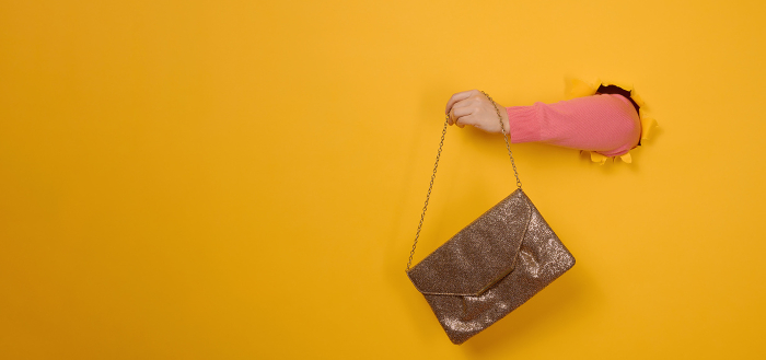 female hand is holding a small golden clutch bag with a metal ch female hand is holding a small golden clutch bag with a metal chain on a yellow background. Part of the body sticking out of a torn hole in a paper background. Copy space
