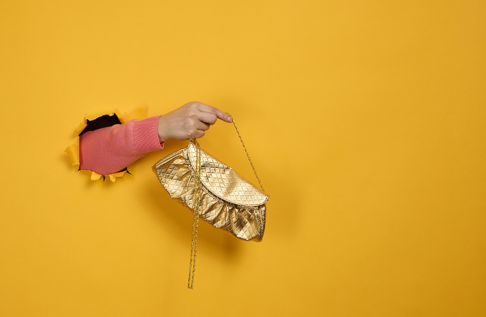 female hand is holding a small golden clutch bag with a metal ch female hand is holding a small golden clutch bag with a metal chain on a yellow background. Part of the body sticking out of a torn hole in a paper background