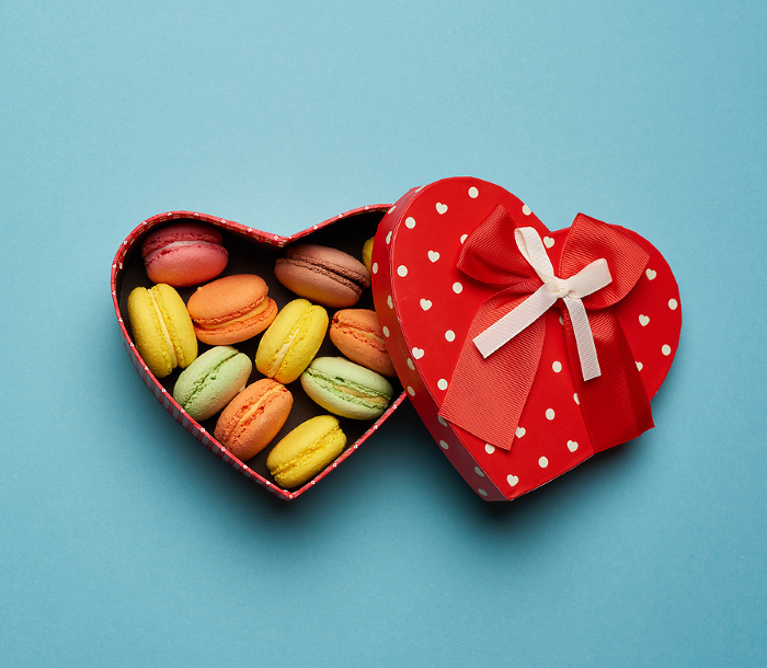 round baked multicolored macarons lie in a red cardboard box in the shape of a heart on a blue background, top view round baked multicolored macarons lie in a red cardboard box in the shape of a heart on a blue background, top view
