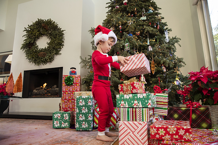 child Boy in stacking up wrapped Christmas presents in front of tree in Santa pajamas.