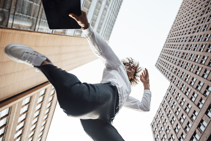 Excited businessman holding digital tablet jumping amidst office buildings, Photo by Gustafsson