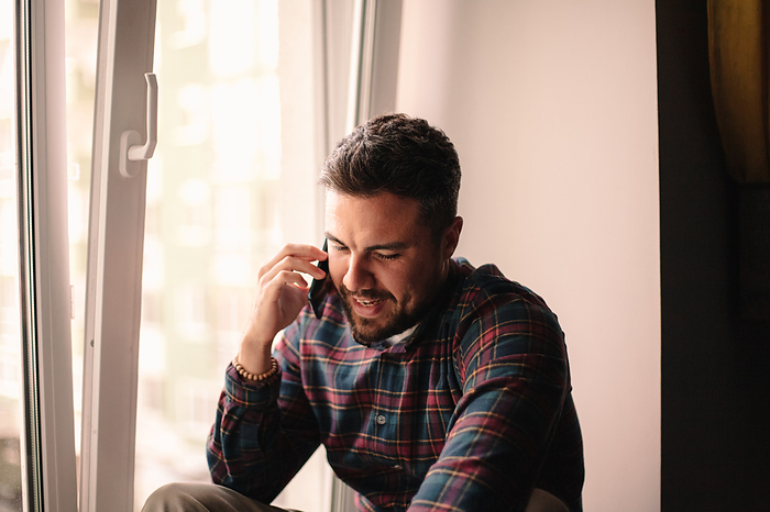 Happy man talking on smart phone sitting by window at home, New York, New York, United States