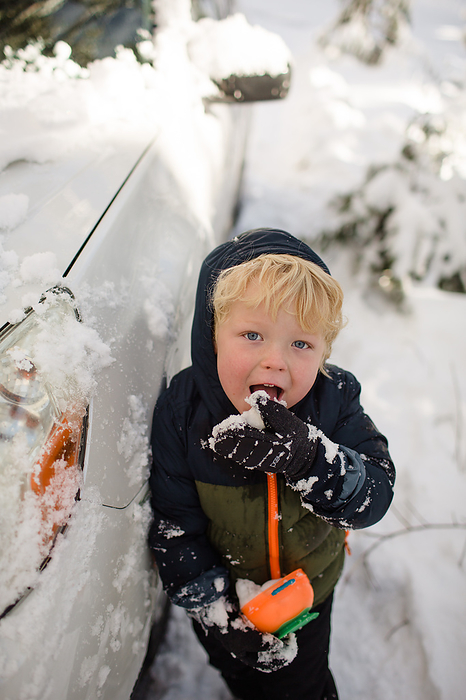 Three Year Old Boy Standing Next to Car Eating Snow