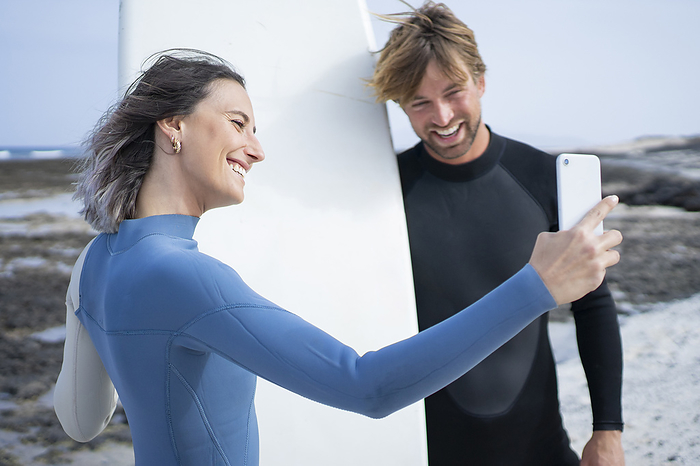 Happy couple in wetsuit  taking selfie with surfboard at beach