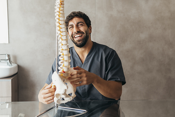 Happy young physiotherapist with human spine model at medical clinic