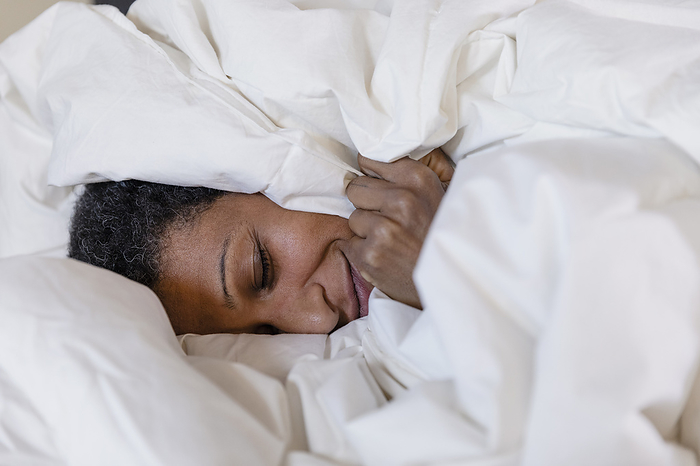 Smiling woman lying on bed covered in white duvet at home