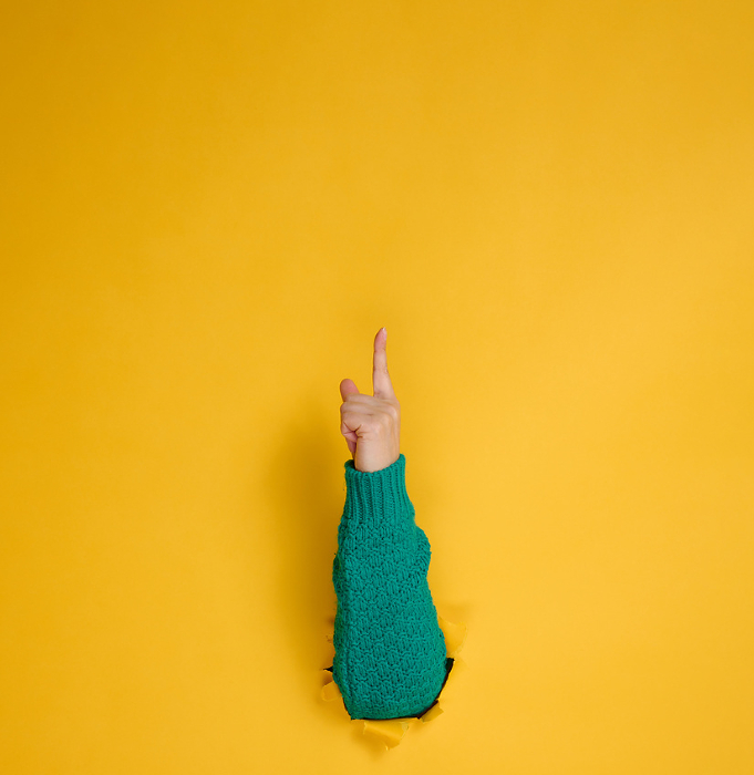 female hand sticks out of a torn hole in a yellow paper background, part of the body points with the index finger to top. Copy space female hand sticks out of a torn hole in a yellow paper background, part of the body points with the index finger to top. Copy space
