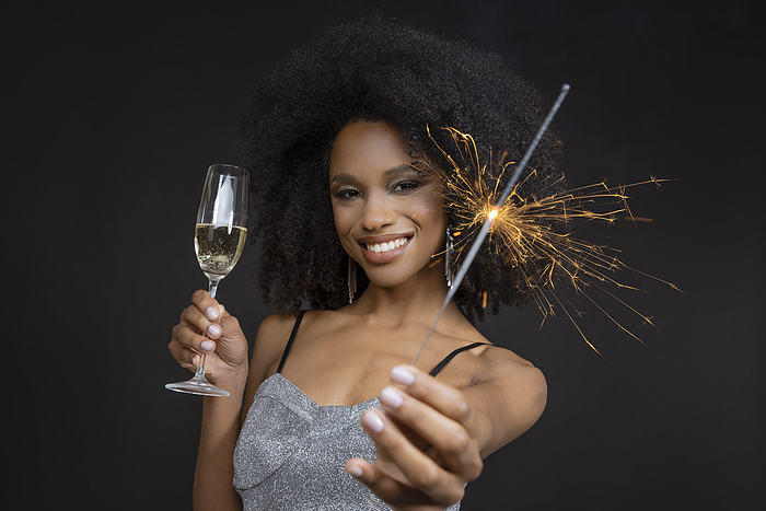 Young black woman with silver party dress holding champagne glass and sparkler Smiling young woman holding champagne flute and sparkler against black background