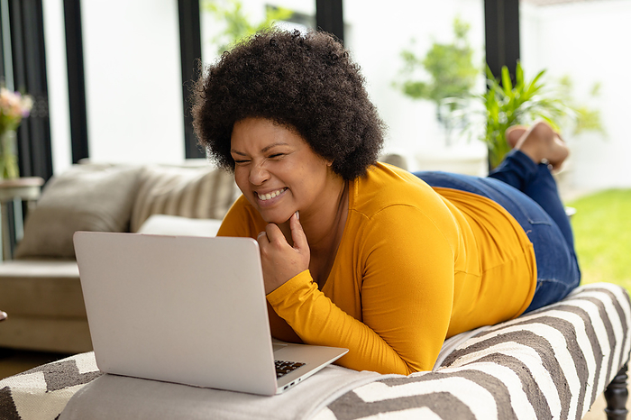 Smiling african american mid adult woman using laptop while lying on couch at home. unaltered, video call, wireless technology, relaxation, lifestyle and domestic life concept.