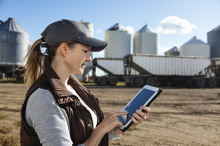 Mature woman farmer working on a tablet computer and standing in front of grain bins at her grainery; Alcomdale, Alberta, Canada
, Photo by LJM Photo / Design Pics