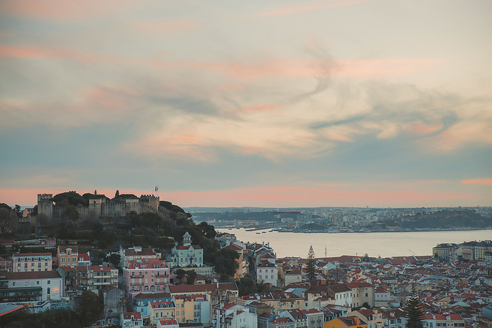 Overview of the Old Town of Portugal's capital city of Lisbon with its pastel colored buildings along the Tagus River and St George's Castle (Castelo de Sao Jorge) on the hilltop in the background at dusk; Lisbon, Portugal, Photo by O'Neil Castro / Design Pics