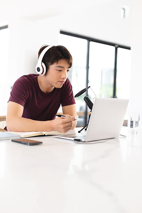 masculine gender Asian teenage boy with microphone on table wearing headphones using laptop while podcasting at home. copy space, unaltered, wireless technology, passion and aspiration concept.