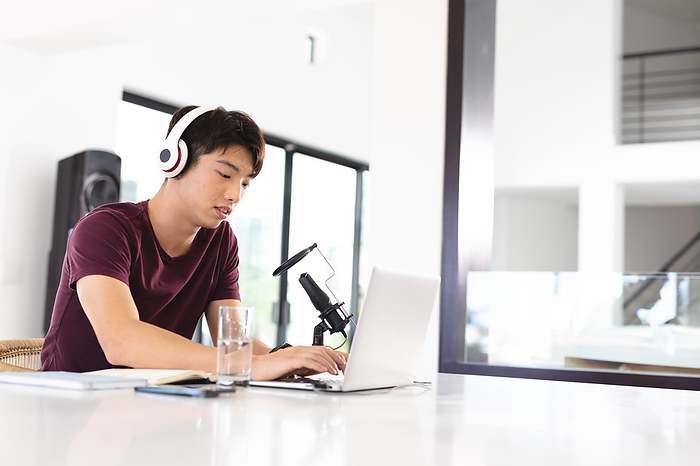 masculine gender Asian teenage boy wearing headphones with microphone on table using laptop while podcasting at home. copy space, unaltered, wireless technology, passion and aspiration concept.