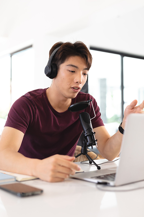 masculine gender Asian teenage boy wearing headphones podcasting over microphone and laptop on table at home. copy space, unaltered, wireless technology, passion and aspiration concept.