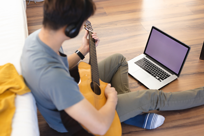 masculine gender High angle view of asian teenage boy playing guitar while learning through laptop on hardwood floor. copy space, wireless technology, unaltered, music, hobbies, lifestyle and home concept.