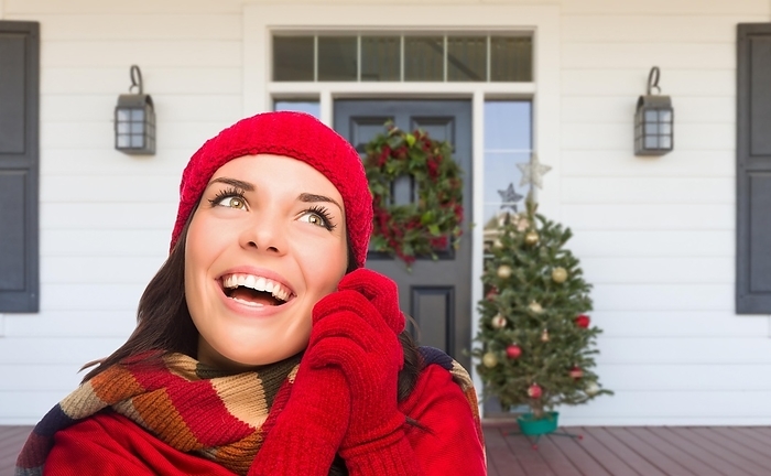 Young girl wearing scarf, red cap and mittens standing on christmas decorated front porch