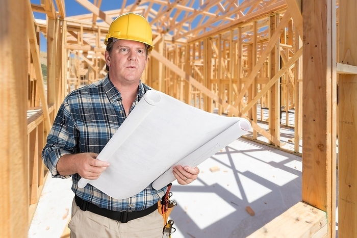 Male contractor with house plans wearing hard hat inside new house construction framing
