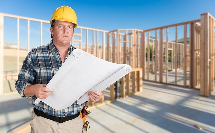 Male contractor with house plans wearing hard hat inside new house construction framing