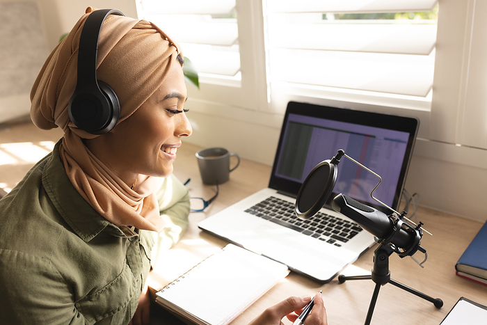 Smiling biracial young woman in hijab recording podcast through condenser microphone at home studio. Unaltered, communication, internet, blogger, broadcasting, influencer, host, podcasting.