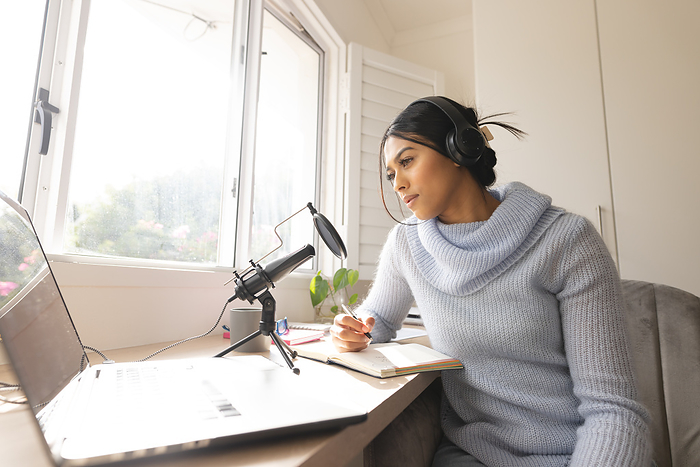 Biracial young woman recording podcast through microphone by window at home studio. Unaltered, lifestyle, communication, internet, blogger, broadcasting, influencer, host, podcasting.