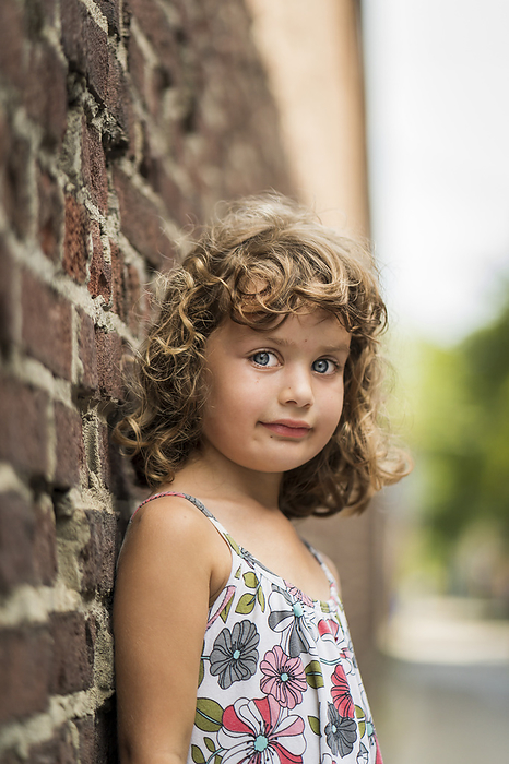 Portrait of a young girl with curly blond hair and blue eyes standing against a brick wall; Toronto, Ontario, Canada, Photo by Ian Taylor / Design Pics
