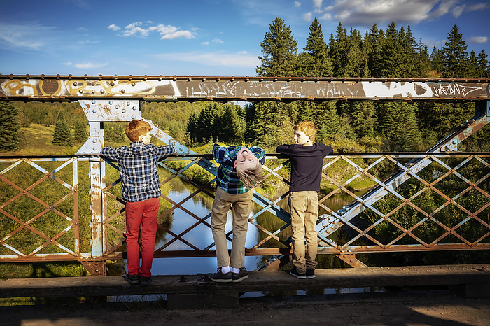 Three boys look out from a park bridge to a creek below, one boy in the middle bending over backwards to look at the camera in a silly pose; Edmonton, Alberta, Canada, Photo by LJM Photo / Design Pics