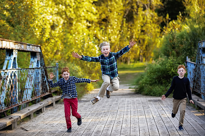 Three boys running across a bridge in a park in autumn, with the boy in the middle leaping high into the air; Edmonton, Alberta, Canada, Photo by LJM Photo / Design Pics