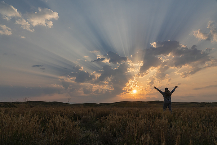 Corpuscular rays during sunrise over the prairies of Saskatchewan. Woman standing and rejoicing in the beauty of a new day; Saskatchewan, Canada, Photo by Robert Postma / Design Pics