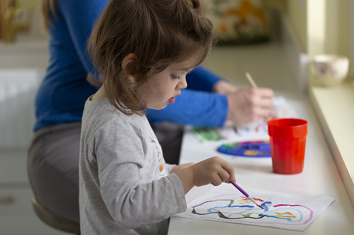 Young girl painting with water colours at home with her mother painting beside her in the background; British Columbia, Canada, Photo by Lorna Rande / Design Pics