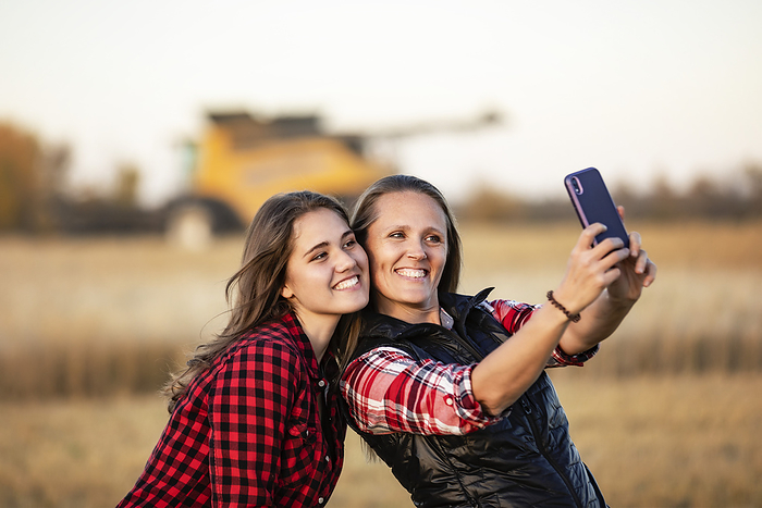 Two farm women posing for a self-portrait on a smart phone at sunset during harvest with a combine working in the background; Alcomdale, Alberta, Canada, Photo by LJM Photo / Design Pics
