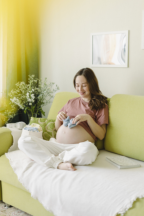 beautiful pregnant Bashkir woman resting at home, sleep, meditation, stretching, drinking regime, self massage, online shopping, video calls Happy woman with baby booties sitting on sofa at home