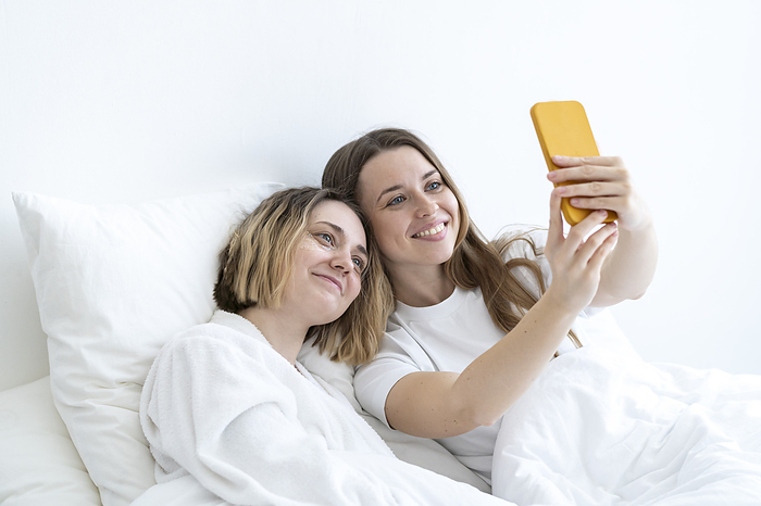 Self care day Happy young friends taking selfie through smart phone on bed at home