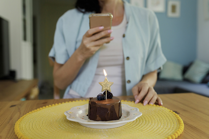 Woman photographing birthday cake with burning star shape candle through mobile phone