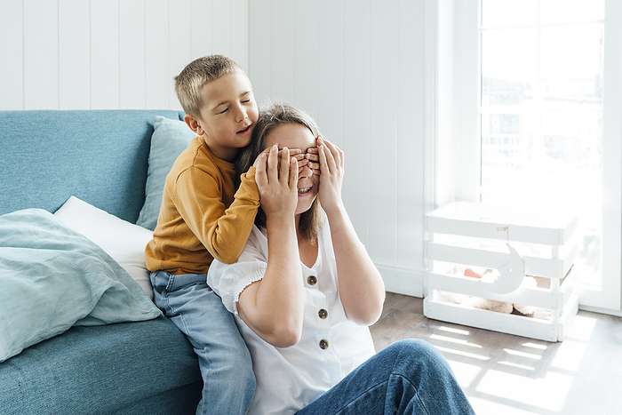 Boy covering eyes of mother sitting by sofa at home
