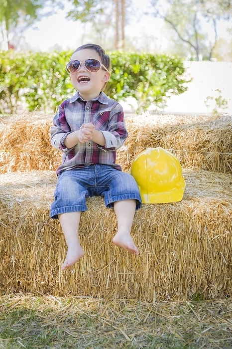 Cute young mixed-race boy laughing with sunglasses and hard hat outside sitting on hay bale, Photo by Andy Dean
