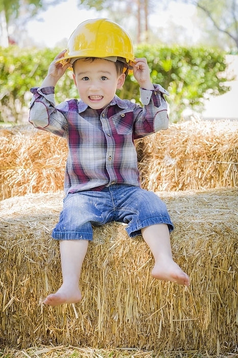 Cute young mixed-race boy laughing with hard hat outside sitting on hay bale, Photo by Andy Dean