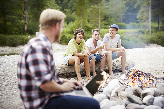 Happy friends looking at businessman with laptop in front of campfire