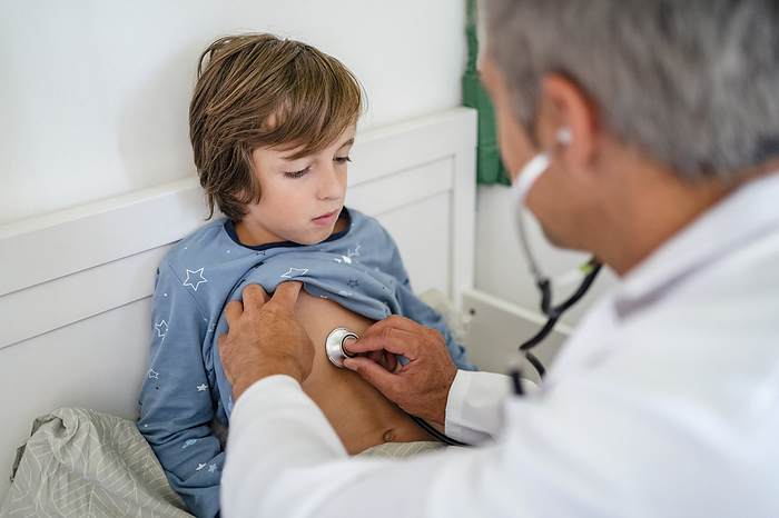 Doctor with stethoscope examining boy in bed at home