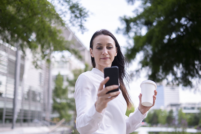 Mature businesswoman using smart phone and holding reusable cup, Photo by ANTHONY PHOTOGRAPHY